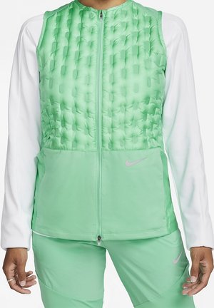 Osoba nosząca jasnozieloną pikowaną kamizelkę na białej koszuli z długim rękawem oraz pasujące jasnozielone spodnie, logo Nike widoczne na kamizelce i spodniach.