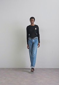 Un model merge cu încredere îmbrăcat într-un top negru cu mânecă lungă și un broșă, purtând blugi albaștri cu talie înaltă, pe un fundal gri simplu.