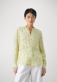Gul langermet bluse med et teksturert mønster av svarte squigglier, med en liten krage og en knappelukking foran.