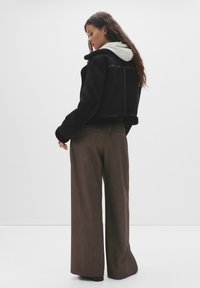 Donna con lunghi capelli che indossa un giubbotto di shearling nero su una felpa grigia e pantaloni marroni larghi, in piedi su uno sfondo bianco.