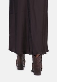 De onderkant van een persoon die een lange donkere rok en bruine leren enkellaarsjes draagt met riemen en klinknagels.