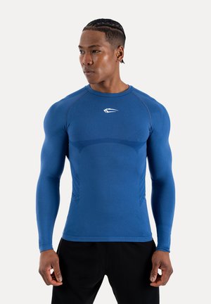 Atletisk mand med fletninger iført en tætsiddende blå langærmet skjorte og sorte bukser, der kigger mod højre.