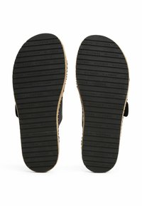 Semelles en caoutchouc noir avec une texture antidérapante portant un nom de marque en relief. Les bords sont finis avec un matériau de couleur claire.