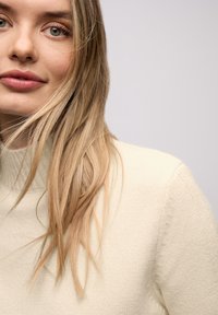 Jasnobeżowy sweter z wysokim kołnierzem, wykonany z miękkiego dzianinowego materiału. Długie pasma włosów opadające na twarz podkreślają czysty design.