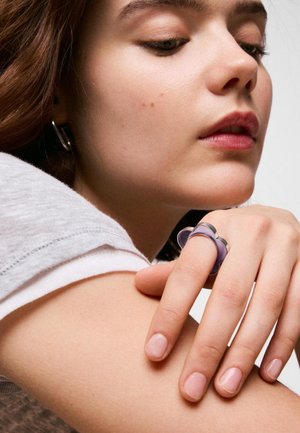 Joven mujer con los ojos cerrados apoyando la barbilla en su brazo, llevando un gran anillo morado y una camisa blanca de manga corta.