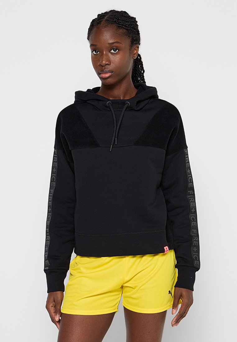 Schwarzer Kapuzenpullover mit Kapuze, gerippten Bündchen und einem strukturierten Vorderteil. Verfügt über ein gedrucktes Logo auf den Ärmeln und ein pinkes Etikett am Saum. Kombiniert mit gelben Shorts.