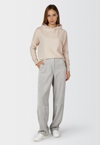 Woman standing wearing a light beige hooded sweater, gray wide-leg pants with subtle stripes, and white sneakers, hands in pockets.