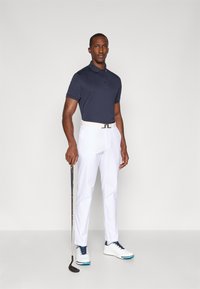 Námořnické polokošile, bílé kalhoty a bílé golfové boty s modrými akcenty. Drží golfovou hůl a stojí na neutrálním pozadí.