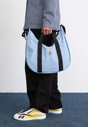 Sac hobo bleu clair avec des poignées noires, comportant un petit patch logo jaune et blanc. Texture lisse, forme arrondie et design spacieux.