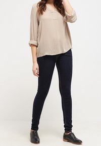 Blouse beige ample avec manches 3/4 et col en V ; jean skinny bleu foncé ; chaussures noires à lacets. Tenue simple et décontractée.