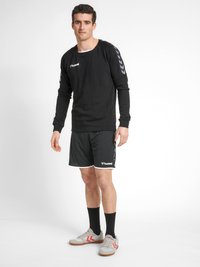 Svart långärmad tröja med rund hals och vitt logotyp, i kombination med svarta sportshorts med vit kant, samt grå sportskor.
