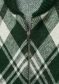Pull en maille à motif vert et blanc avec une fermeture éclair métallique visible, présentant un motif pied-de-poule et un col côtelé.