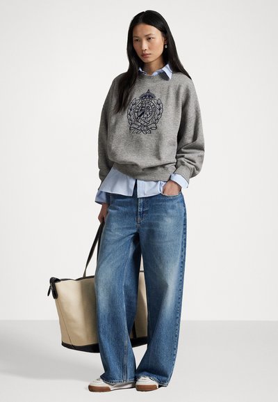 Felpa grigia con emblema ricamato, indossata sopra una camicia colletto blu chiaro. Abbinata a jeans a gamba larga blu e una borsa a tracolla beige.