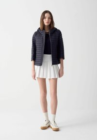 Korte marineblauwe gewatteerde jas met een rits aan de voorkant en kap, gedragen over een strakke donkere top en een witte geplooide rok, gecombineerd met witte sneakers.