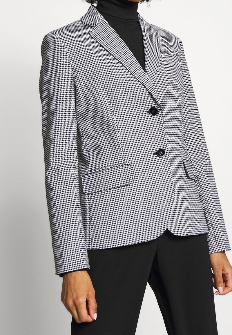 Mustapohjainen houndstooth-kuvioinen bleiseri, jossa on lovet, kaksi nappia ja etutaskut. Päällä musta poolopaita.