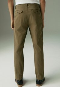 Pantalons en coton marron avec une coupe droite, dotés de deux poches arrière et d'une texture lisse. Portés avec des chaussures noires présentant des semelles contrastées.