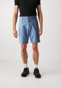 Lyseblå joggeshorts laget av stoff, med brettet kant, to frontlommer og knappelukking. Båret med en svart T-skjorte og sneakers.