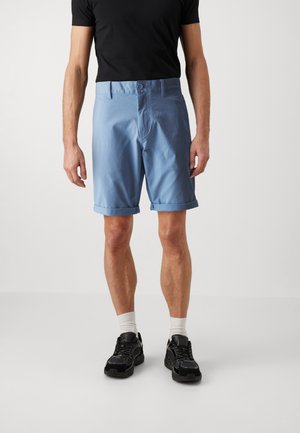 Pantalones cortos informales de color azul claro hechos de tela, con un dobladillo doblado, dos bolsillos delanteros y un cierre de botón. Llevados con una camiseta negra y zapatillas.