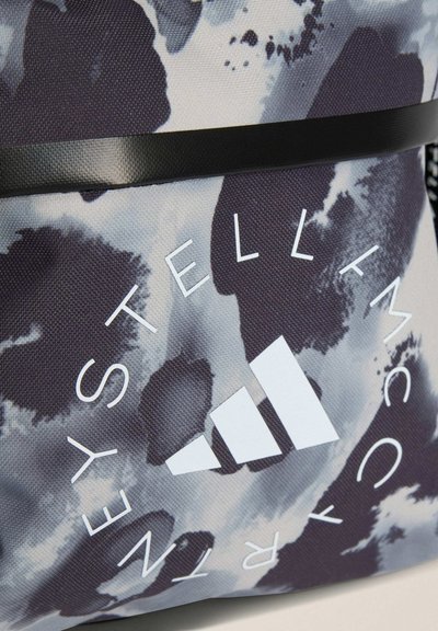 Zbliżenie na szaro-czarną tkaninę w kamuflażowy wzór z logo Adidas i napisem "Stella McCartney" w białym kółku.