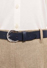 Cintura di pelle blu navy con fibbia rotonda argentata allacciata sopra pantaloni di lino beige e una camicia bianca con bottoni.