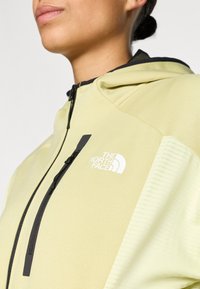 Jachetă galben deschis cu glugă, fermoar negru și logo. Dispune de panouri netede și texturate, culori contrastante și un design alungit.