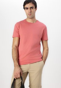 Pink crew neck t-shirt made of soft fabric, short sleeves, paired with beige shorts. The figure holds a black bag, standing against a white background.