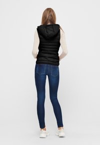 Černá p�éřová vesta s kapucí, prošívaná textura. V kombinaci s tmavě modrými skinny džínami a bílými sportovními botami. Pohled zezadu.