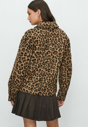 Vrouw met lang donker haar in een panterprint jasje en een zwarte geplooide mini rok, zijwaarts kijkend tegen een effen achtergrond.