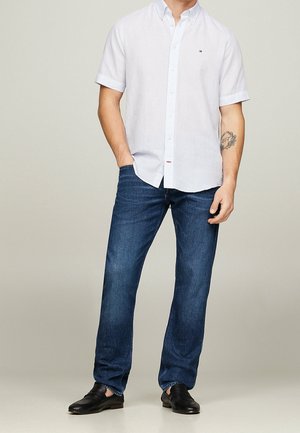 Lichtblauw overhemd met korte mouwen en knopen, spijkerbroek en zwarte schoenen. Het overhemd heeft een kraag en een klein logo; de spijkerbroek heeft een klassieke pasvorm.