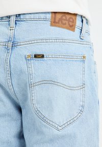 Vaaleansiniset denim-farkut ruskealla nahkatarroilla, jossa on "Lee" vyötäröllä ja pieni musta merkki takataskun lähellä.