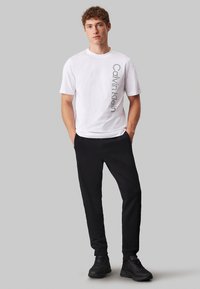 T-shirt bianco in cotone con il logo "Calvin Klein" stampato verticalmente in nero, abbinato a pantaloni della tuta neri e scarpe da ginnastica nere.