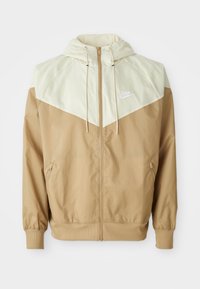 Nike windbreaker jacket featuring a beige body, light tan paneling, hood, front zipper, and elastic cuffs with a small white logo on the chest.