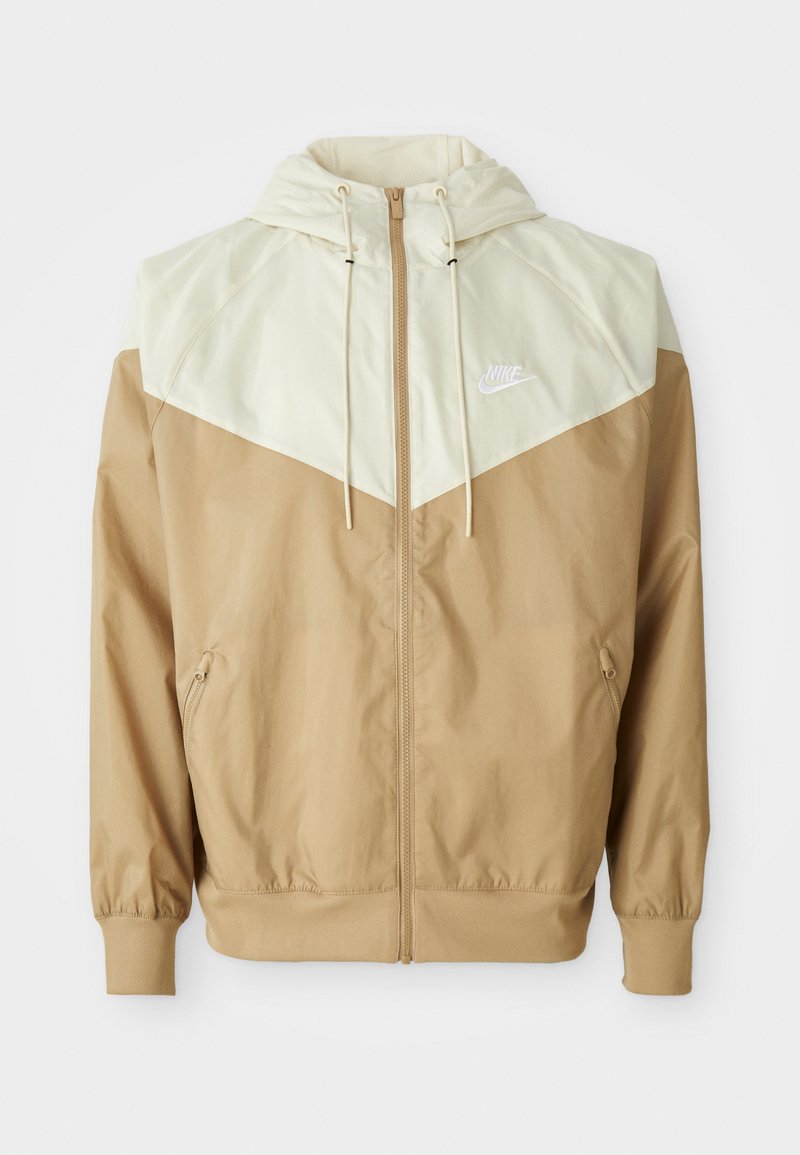 Nike windbreaker jacket featuring a beige body, light tan paneling, hood, front zipper, and elastic cuffs with a small white logo on the chest.