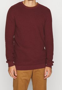 Bordeauxrode geribde trui met lange mouwen, ronde hals, gestructureerd oppervlak en aansluitende boordjes, gecombineerd met lichtbruine broek.