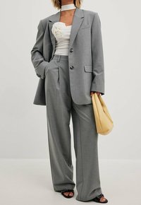 Šedý upravený blazer s odpovídajícími širokými kalhotami, doplněný bílým topem s volány a žlutou kabelkou, s černými botami na otevřenou špičku.