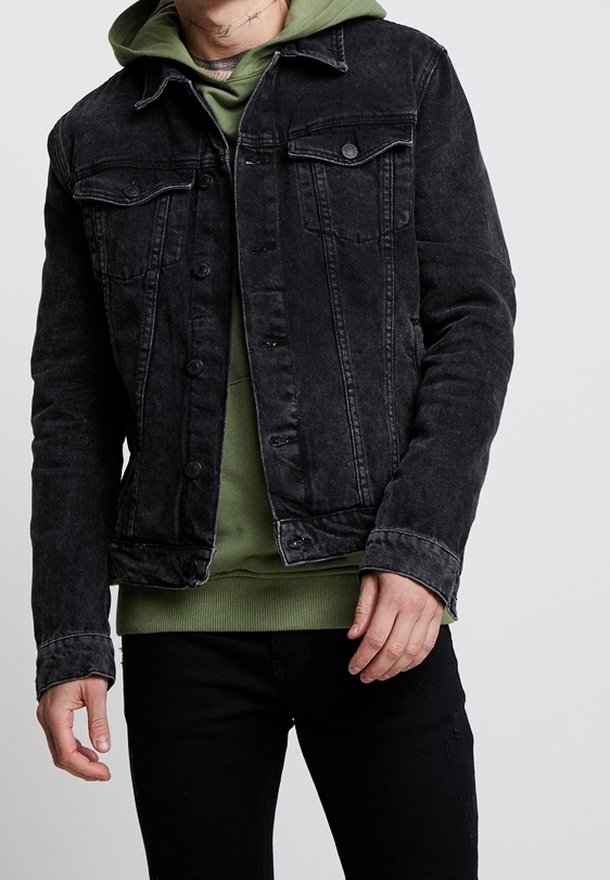 Chaqueta de mezclilla negra con dos bolsillos en el pecho, cierre de botones y costuras negras a juego. Superpuesta sobre una sudadera verde con capucha.