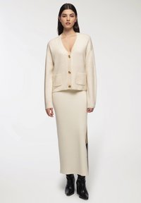 Cardigan beige chiaro con scollo a V, tre bottoni e due tasche anteriori, abbinato a un vestito midi a coste coordinato e stivali neri con tacco.