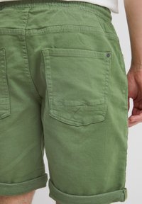 Pantaloncini verdi in cotone con risvolti arrotolati, dotati di due tasche posteriori, una con bottone, e motivo a punto a zigzag sulla patta della tasca.
