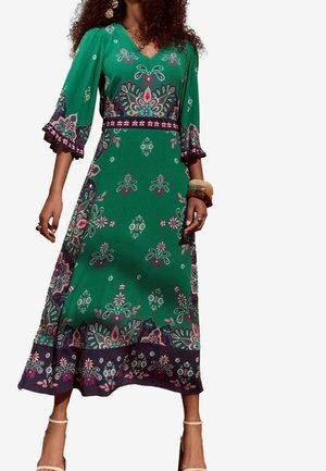 Sukienka maxi z zielonego materiału z misternymi kwiatowymi i geometrycznymi wzorami, z dekoltem w szpic i rozkloszowanymi rękawami z falistymi brzegami.