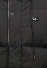 Čierna puffová bunda so zipsom, okrúhlym cvokom, nastaviteľnou kapucňou so šnúrkou a strieborným logom na hrudi.