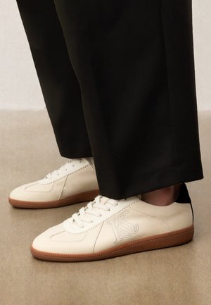 Zapatillas blancas de cuero con suelas de goma y detalle negro en el talón, combinadas con pantalones negros de pierna ancha sobre un suelo beige.