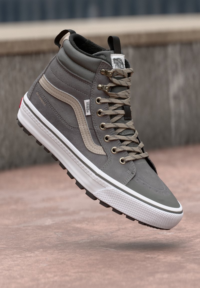 Baskets montantes Vans grises et beige avec lacets, semelle blanche et étiquette imperméable, suspendues au-dessus d'une surface en béton en plein air.