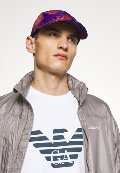 Casquette de baseball avec un motif abstrait multicolore. Le modèle porte une veste zippée légère grise et un t-shirt blanc avec des rayures marines et un logo.