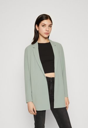 Lichtgroene blazer met lange mouwen, open voorkant en getailleerd ontwerp. Gemaakt over een zwart crop top en gecombineerd met donkere jeans.
