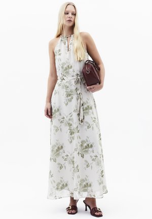 Frau trägt ein weißes, geblümtes Maxikleid mit Neckholder und gebundener Taille, hält eine weinrote Handtasche und trägt passende Sandaletten mit Absatz.