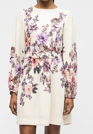 Femme portant une robe blanche à manches longues avec un motif floral dans des tons violets, roses et beiges, avec une ceinture assortie à boucle ronde.