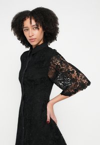 Čierne čipkované šaty s výrazným golierom, zapínaním na gombíky na prednej strane a tromištvrte zvonovými rukávmi s kvetinovými čipkovanými vzormi.