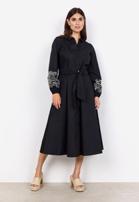 Vestido negro de botones con mangas largas, que presenta bordados florales blancos en las mangas, una cintura ajustada y una falda fluida. Llevado con sandalias de plataforma.