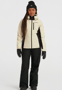 Beige ski-jack met patroon en zwarte panelen, met ritszakken, gecombineerd met zwarte skibroek en handschoenen. Een zwarte gebreide muts maakt de outfit compleet.