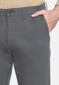 Hombre que lleva pantalones grises con botón y cremallera, mano casualmente en el bolsillo delantero derecho, suéter beige visible en la cintura.